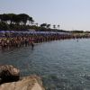 Una spiaggia affollata con persone in fila lungo la riva, ombrelloni blu e alberi sullo sfondo.